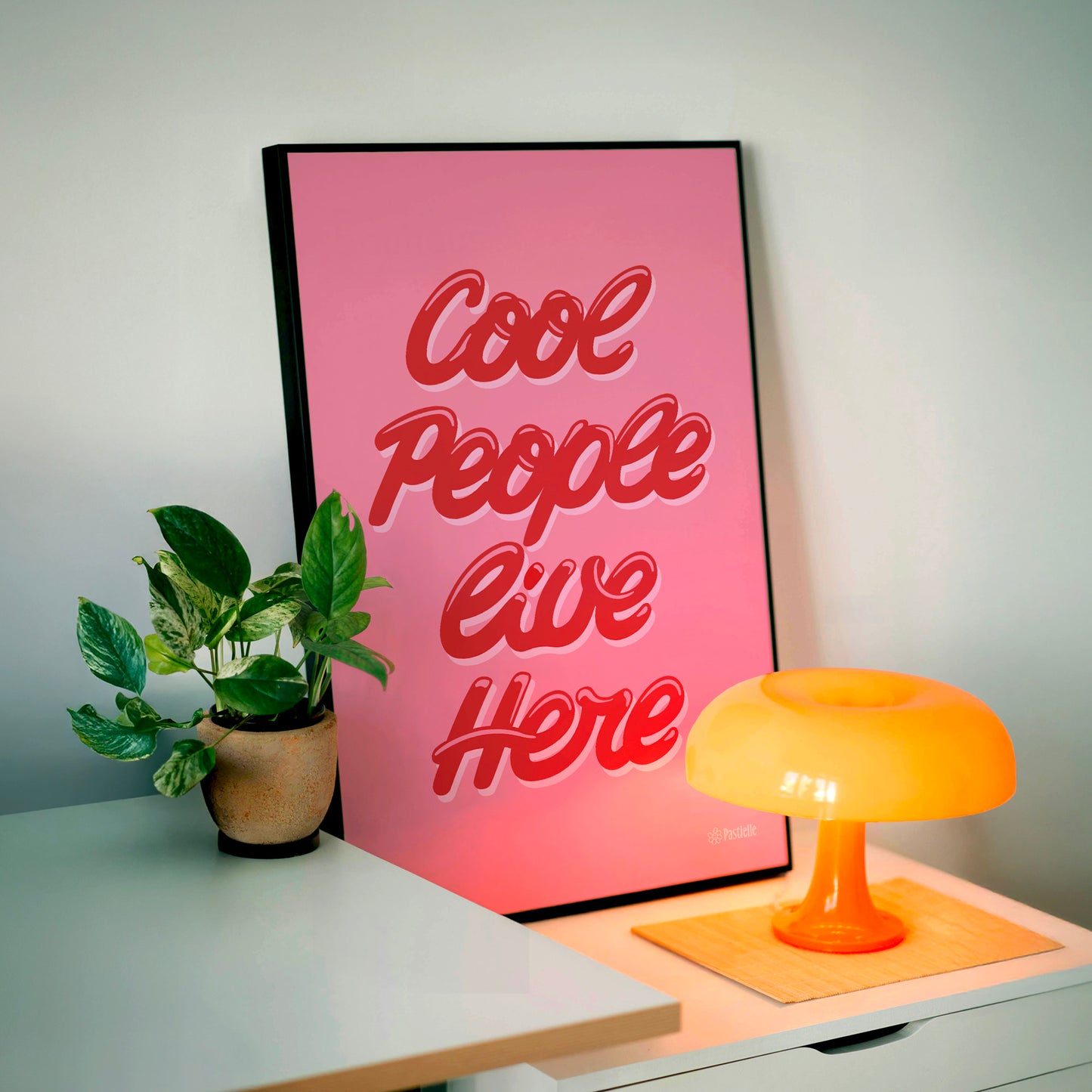 Affiche rétro Pastielle Cool People Live Here – affiche rose posée sur une table avec lampe orange et plante verte