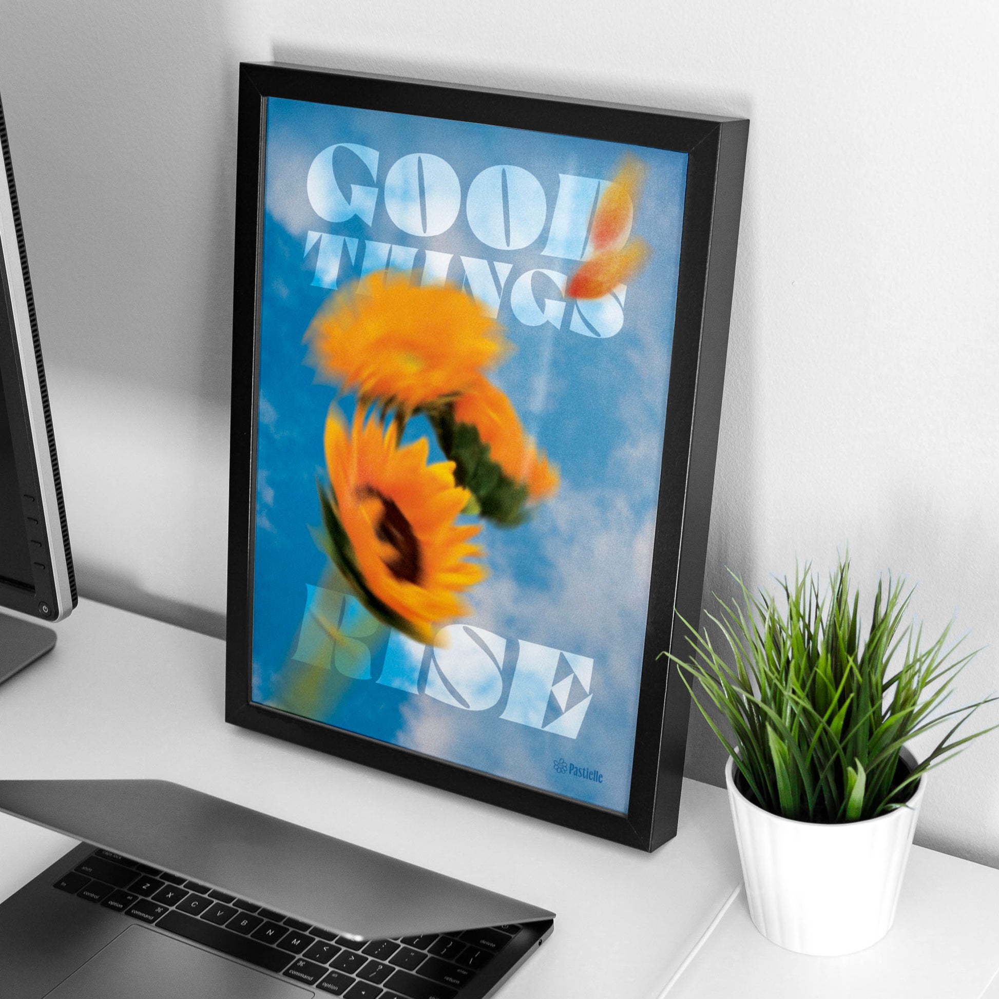 Affiche murale design Pastielle Good things rise – affiche représentant un tournesol devant un ciel bleu, cadre noir posé sur un bureau avec une plante et un ordinateur