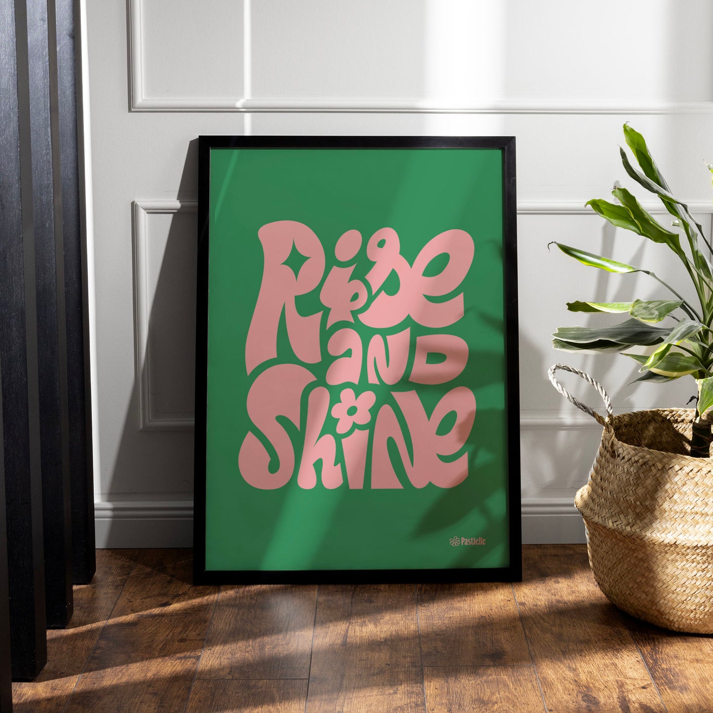 Affiche rise and shine écrit en rose sur fond vert dans un cadre, posée au sol avec une plante à côté
